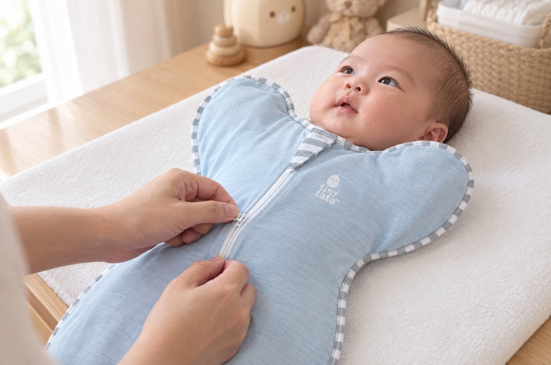 Parent zipping a newborn into a light blue TatoWrap zip swaddle on a changing mat, baby on their back with arms up in the wings