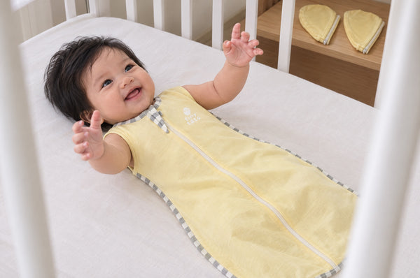 Smiling baby on their back in a cot wearing a yellow TatoWrap+ swaddle with the sleeves removed for arms-out sleep, detached sleeves on a shelf