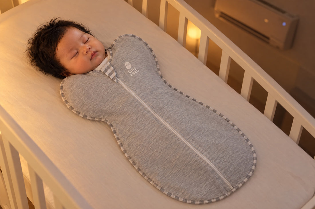Newborn asleep on their back in a cot at night, wearing a grey TatoWrap zip swaddle in a room with the aircond on
