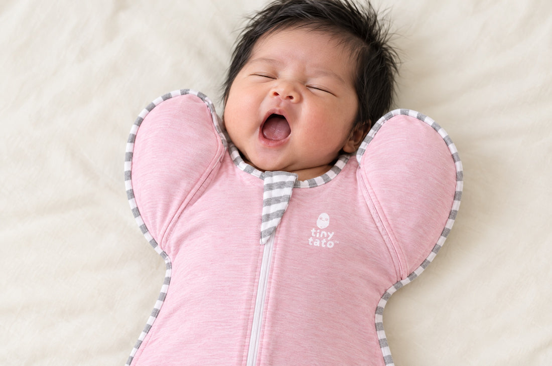 Newborn yawning on their back in a pink TatoWrap zip swaddle with arms up in the wings near the face
