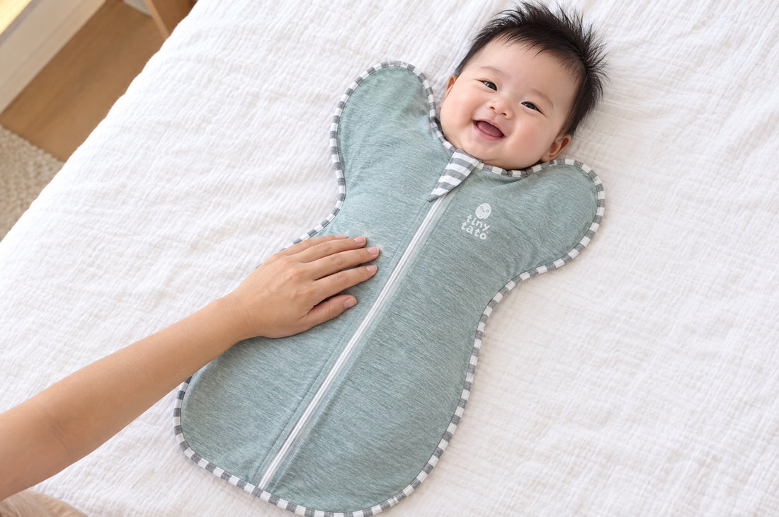 Smiling baby on a bed in a teal TatoWrap zip swaddle with a roomy bottom so the legs can bend at the hips, parent's hand on the tummy