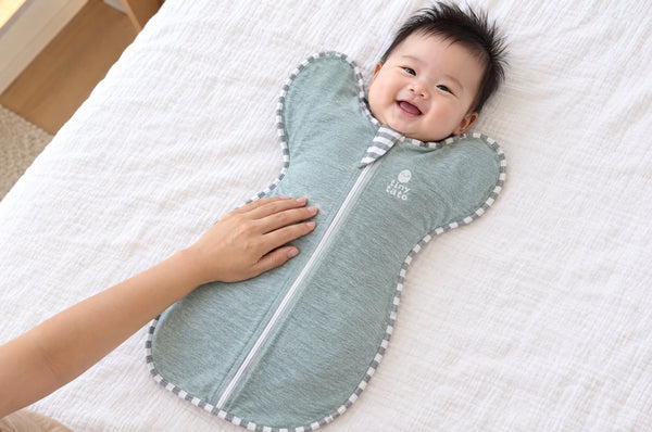 Smiling baby on a bed in a teal TatoWrap zip swaddle with a roomy bottom so the legs can bend at the hips, parent's hand on the tummy