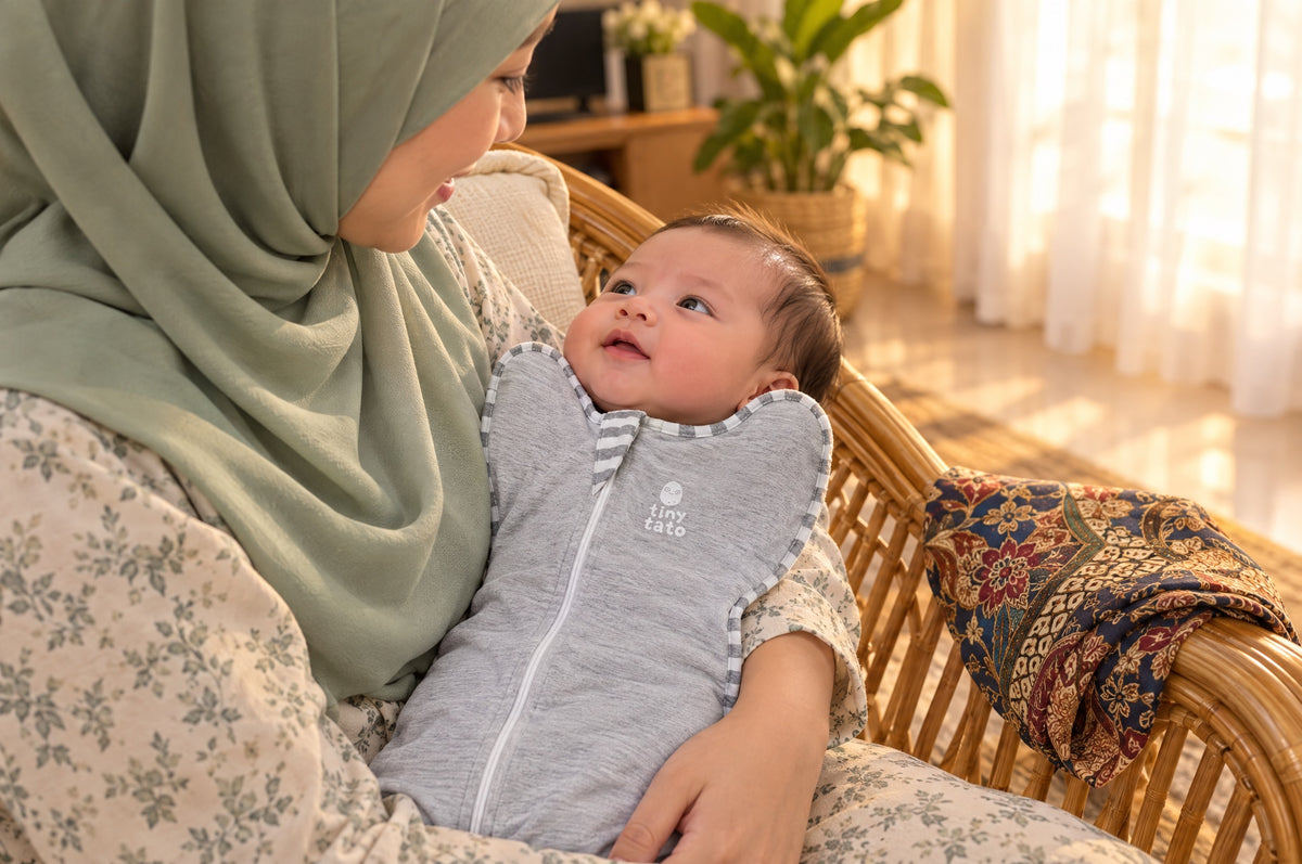 Ibu bertudung mendukung bayi dalam bedung zip TatoWrap warna kelabu di kerusi rotan, dengan kain batik di lengan kerusi