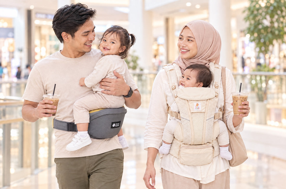 Malaysian dad carrying a toddler on the yellow seat of a TatoHip hip seat carrier beside mum wearing a baby in a TatoSnug structured carrier at a mall