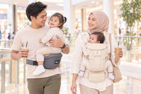 Malaysian dad carrying a toddler on the yellow seat of a TatoHip hip seat carrier beside mum wearing a baby in a TatoSnug structured carrier at a mall