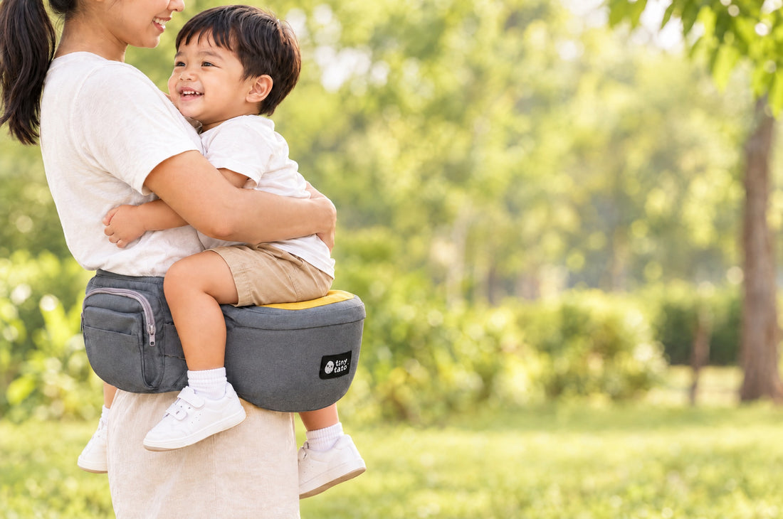 Toddler sitting on the yellow seat of a TatoHip hip seat carrier in the M-position with knees higher than the bottom, parent's arm around them